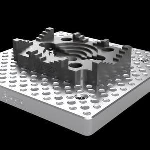 Könnte beinhalten: Ein silberfarbenes Metallteil mit komplexem Design, das eine zentrale kreisförmige Aussparung und abgestufte Kanten aufweist. Es liegt auf einer perforierten Metallplatte. Das Wort "REKER" ist auf der Seite sichtbar.