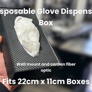 Puede incluir: Un dispensador de pared negro para guantes desechables. Tiene un acabado de fibra de carbono óptica y se adapta a cajas de 22 cm x 11 cm. El texto "Disposable Glove Dispenser Box" está impreso en la parte superior de la caja.