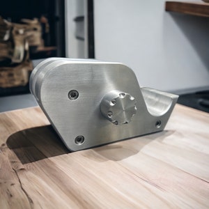 May include: A silver metal phone stand shaped like a whale with a knob on the side. The stand is sitting on a wooden surface.