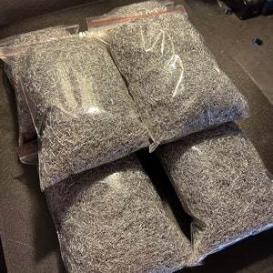 May include: Four clear plastic bags filled with silver metal shavings. The bags are stacked on top of each other on a dark gray surface.