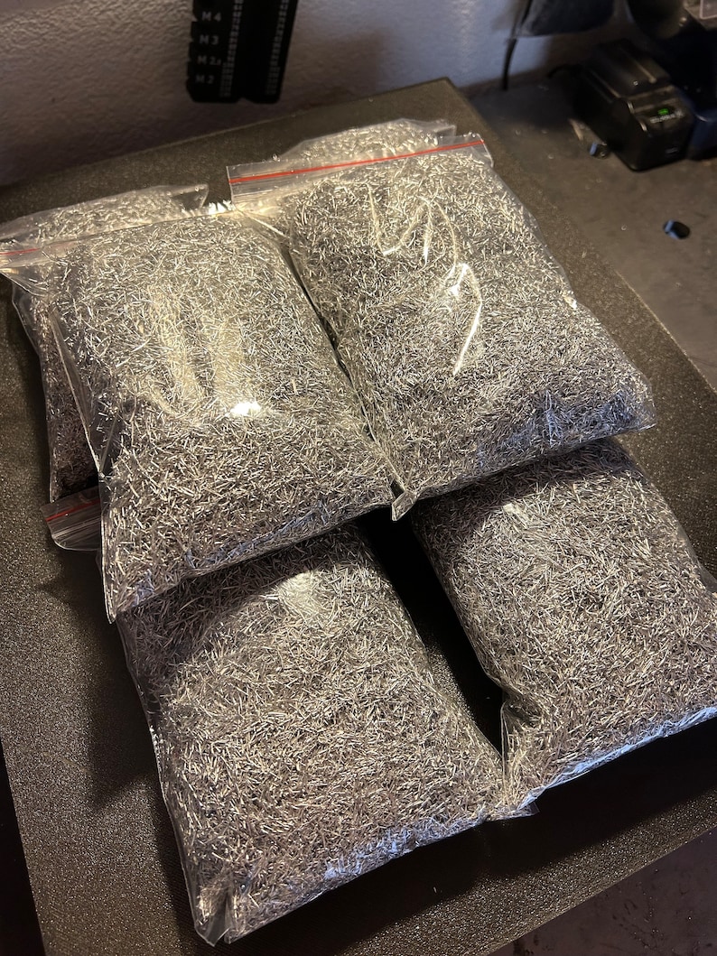 May include: Four clear plastic bags filled with silver metal shavings. The bags are stacked on top of each other on a dark gray surface.
