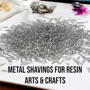 May include: A white plate filled with silver metal shavings. The plate is on a wooden surface. The text "METAL SHAVINGS FOR RESIN ARTS & CRAFTS" is printed below the plate.