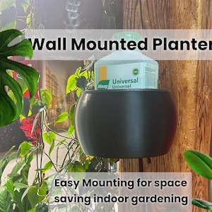 Puede incluir: Un macetero negro de pared con una botella verde de fertilizante para plantas en su interior. El macetero está colgado en una pared de madera. El texto "Wall Mounted Planter" está por encima del macetero y el texto "Easy Mounting for space saving indoor gardening" está por debajo del macetero.