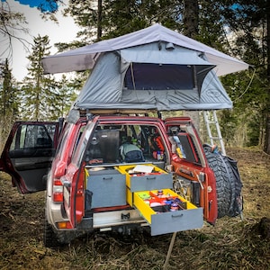May include: A red SUV with a roof top tent and a ladder. The back of the SUV is open and has several storage containers. The containers are labelled with the words "Adventure Ready".