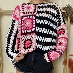May include: A white, black, and pink crocheted sweater with a square pattern. The sweater has long sleeves and a loose fit.