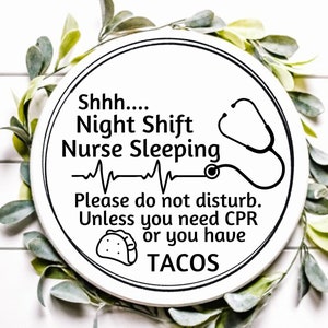 Puede incluir: Un letrero redondo de madera blanca con texto negro y un gráfico de estetoscopio. El letrero dice: "Shhh... Enfermera de turno de noche durmiendo. Por favor, no molestar. A menos que necesites RCP o tengas TACOS."