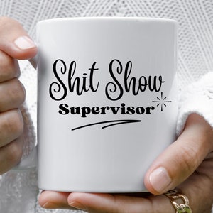 May include: White ceramic coffee mug with black text that reads "Shit Show Supervisor" with a star and a line underneath.