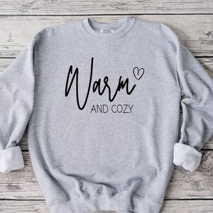 May include: Gray sweatshirt with the text "Warm" in a cursive font and a small heart symbol. The text "and cozy" is written below in a smaller font.