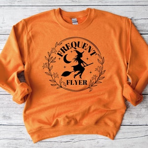以下が含まれることがあります： オレンジ色のスウェットシャツに、ほうきに乗って飛んでいる魔女の黒いグラフィックが描かれており、円の中に「Frequent Flyer」という文字が書かれています。