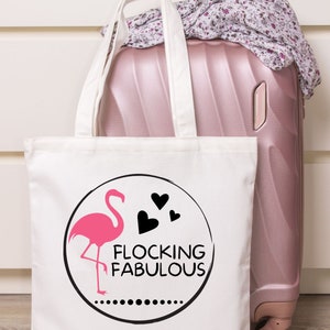 May include: White canvas tote bag featuring a pink flamingo and black hearts within a circle. The text "Flocking Fabulous" is displayed beneath the flamingo. The bag is positioned on top of a pink suitcase.