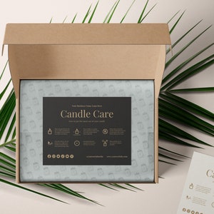 May include: A brown cardboard box with the lid open, revealing a white tissue paper with a black and gold label that says "Candle Care". The label has instructions on how to get the most out of your candles. The box is sitting on a light beige surface with a green palm leaf.