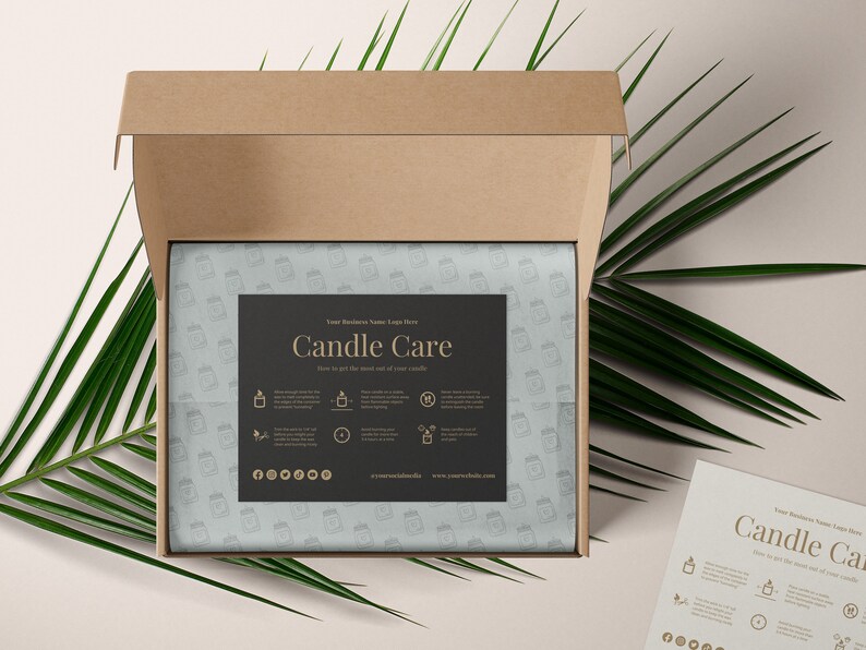 May include: A brown cardboard box with the lid open, revealing a white tissue paper with a black and gold label that says "Candle Care". The label has instructions on how to get the most out of your candles. The box is sitting on a light beige surface with a green palm leaf.