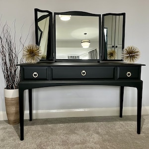 May include: A black vanity table with three mirrors and three drawers. The table has a simple design with gold-coloured handles.