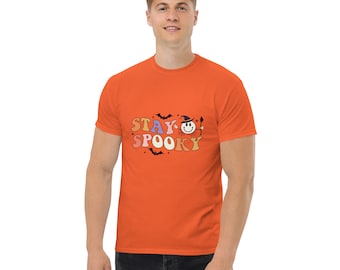 May include: Orange short-sleeved t-shirt with the text "Stay Spooky" in a retro font. The text is in shades of pink, purple, orange, and yellow. A smiling cartoon face with a witch's hat is in the middle of the text.