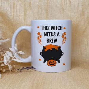 Op de afbeelding: Witte keramische mok met een zwarte ketel, een oranje pompoen en de tekst "THIS WITCH NEEDS A BREW". De mok heeft een gebogen handvat en is versierd met oranje bellen. Perfect voor Halloween.