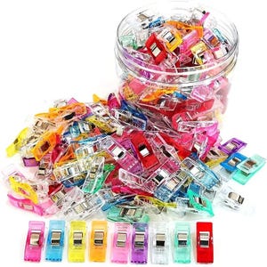 May include: A large collection of small, multi-coloured plastic sewing clips with metal springs. The clips are translucent in shades of red, blue, yellow, green, orange, and pink. Many clips are inside a clear plastic jar, while others are scattered.