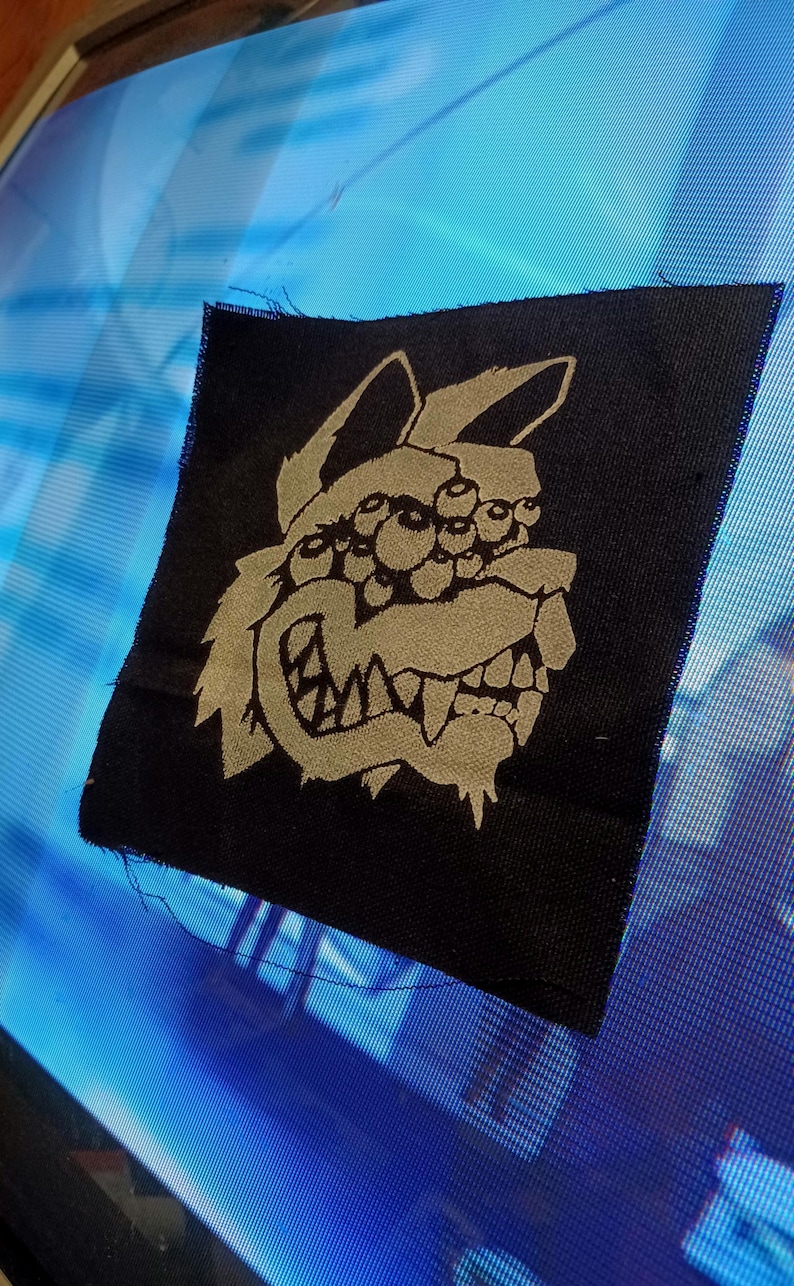 Pode incluir: Remendo de tecido preto com uma imagem bordada branca de uma criatura com m&uacute;ltiplos olhos e uma grande boca.