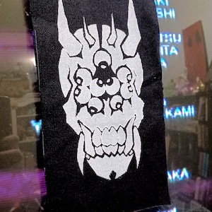May include: Black rectangular patch with a white Oni mask design. The mask features horns, multiple eyes, and a wide mouth. The patch is likely for clothing or accessories.