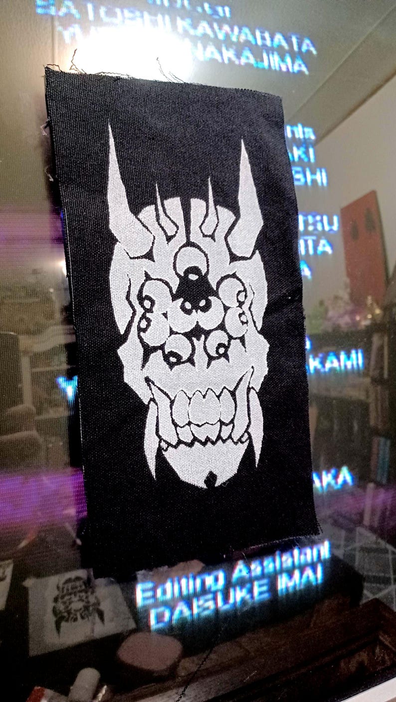 May include: Black rectangular patch with a white Oni mask design. The mask features horns, multiple eyes, and a wide mouth. The patch is likely for clothing or accessories.