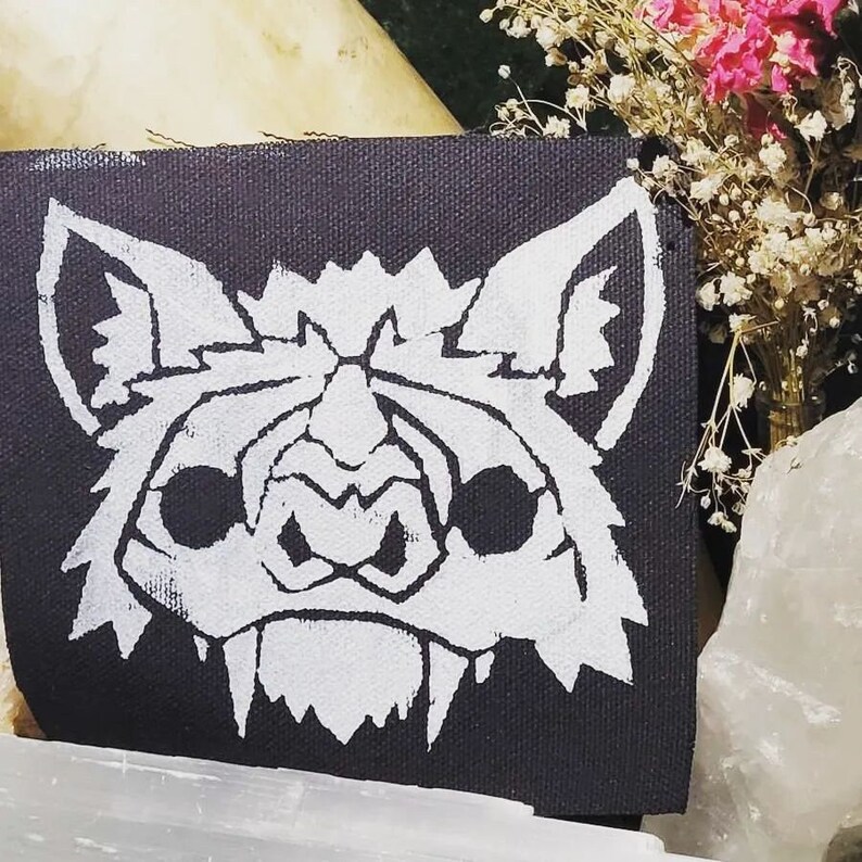 May include: A black fabric patch featuring a white silhouette of a bat's face. The bat has large, round eyes and sharp teeth.