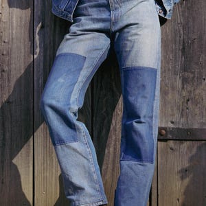 May include: High-waisted blue denim jeans featuring rectangular patches of a darker blue fabric on the front of each thigh. The jeans are paired with a brown leather belt with a large metal buckle, a white shirt, and a blue denim jacket.