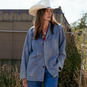 May include: A light blue denim jacket with a button-down front, a collar, and two large patch pockets on the lower front. The jacket is paired with a white cowboy hat and bright blue trousers featuring visible stitching and panel details.