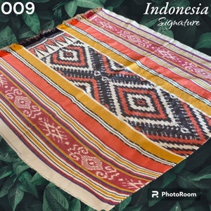 Könnte beinhalten: Eine farbenfrohe gewebte Wolldecke mit einem geometrischen Muster in Rot-, Gelb-, Schwarz- und Weißtönen. Die Decke hat einen Fransenrand und ist mit "Indonesia Signature" beschriftet.