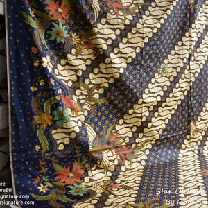 Puede incluir: Una tela batik azul y dorada con un patrón repetitivo de estrellas y remolinos. La tela está doblada por la mitad y drapeada sobre una pila de madera.