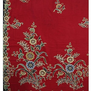 Op de afbeelding: Een levendig rood textiel met een bloemenpatroon. Het ontwerp bevat gestileerde bloemen in tinten blauw, geel en groen, met donkergroene bladeren en stelen. De randen zijn afgezet met een zwart-wit bloemenpatroon.