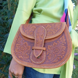 Könnte beinhalten: Eine braune Ledertasche mit aufwendigen floralen Schnitzereien und einem Klappverschluss. Die Tasche hat weiße Nähte an den Rändern und eine Metallschnalle.