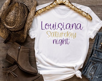 May include: White t-shirt with the text "Louisiana Saturday night" in purple and gold glitter lettering.