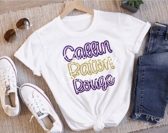 May include: White t-shirt with the text "Geaux Baton Rouge" in purple and gold glitter lettering.