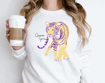 May include: White sweatshirt featuring a yellow and purple tiger graphic with the text "Geaux Tigers." The sweatshirt is being held by a person holding a white coffee cup with a brown sleeve.
