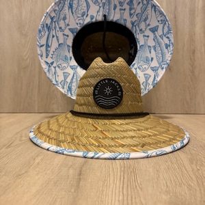 May include: A straw sun hat with a wide brim and a black cord. The brim is trimmed with a white border featuring a blue fish pattern. The hat has a black patch with the text "LITTLE JACKS" and a sun graphic.
