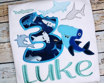 May include: White t-shirt with a blue and teal shark design. The number "3" is filled with shark illustrations and the name "LUKE" is embroidered in teal script. Features a hammerhead shark.