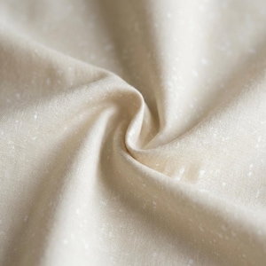 May include: Close-up of a beige fabric with a subtle speckled pattern. The fabric is gathered and folded, creating soft shadows and highlighting the texture. The neutral color and delicate design suggest a natural textile.