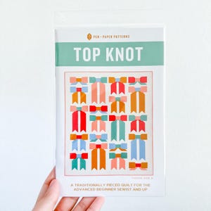 May include: A sewing pattern titled "TOP KNOT" in a clear plastic sleeve. The pattern features a colorful quilt design with bow shapes in red, orange, pink, blue, and green. The text "PEN • PAPER PATTERNS" is at the top.