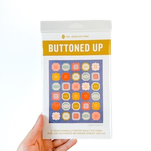 May include: A package for a "Buttoned Up" large throw quilt pattern. The package features a blue background with colorful button designs. The text "Buttoned Up" is in large, gold letters. The package is held by a hand.