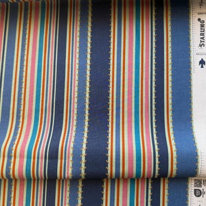 May include: Close-up of a fabric with vertical stripes in shades of blue, pink, orange, and cream. The fabric has a stitched design along the blue stripes. The fabric is labeled "100% Premium Cotton".