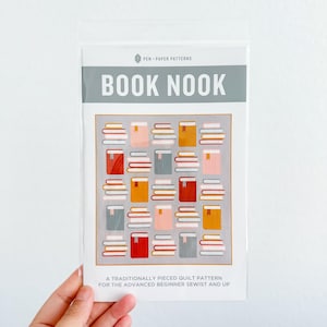 May include: A clear plastic package containing a quilt pattern. The pattern features a gray background with a grid of colorful, stacked books. The text "BOOK NOOK" is displayed prominently, with the words "PEN - PAPER PATTERNS" above and pattern details below.