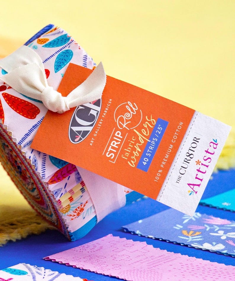 May include: A fabric strip roll tied with a cream ribbon. The roll features colorful patterned fabric with red, blue, and white designs. An orange tag reads "STRIP Roll wonders" and "40 STRIPS / 2.5"" with the brand name "Art Gallery Fabrics".