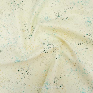 May include: Cream-colored fabric with a speckled pattern. The fabric features small, irregular spots in teal, dark green, and gold. The fabric is slightly wrinkled, suggesting it is a soft material.