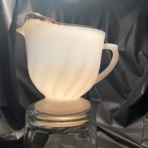 May include: White milk glass creamer with a gold rim and a swirl design. The creamer is sitting on a clear glass tumbler.