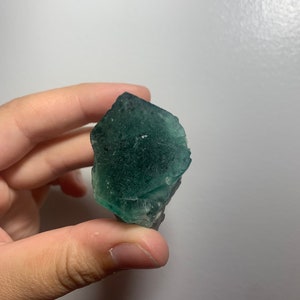 May include: A green fluorite crystal with a rough, natural texture. The crystal is translucent and has a slight shimmer.