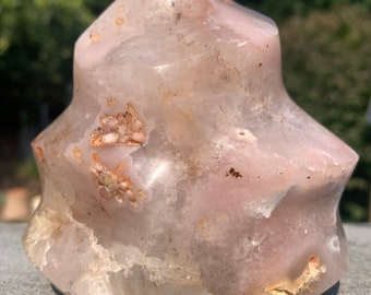 May include: A polished, freeform crystal sculpture with a light pink and white color palette. The stone has a unique, flame-like shape with rounded edges and visible natural inclusions. The crystal sits on a gray surface, with a blurred green background.