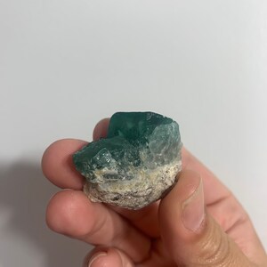 May include: A green fluorite crystal with a rough, natural texture. The crystal is a deep green colour and has a slightly cloudy appearance.