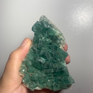 May include: A cluster of green fluorite crystals with a rough, natural texture. The crystals are arranged in a pyramid shape and have a translucent appearance.