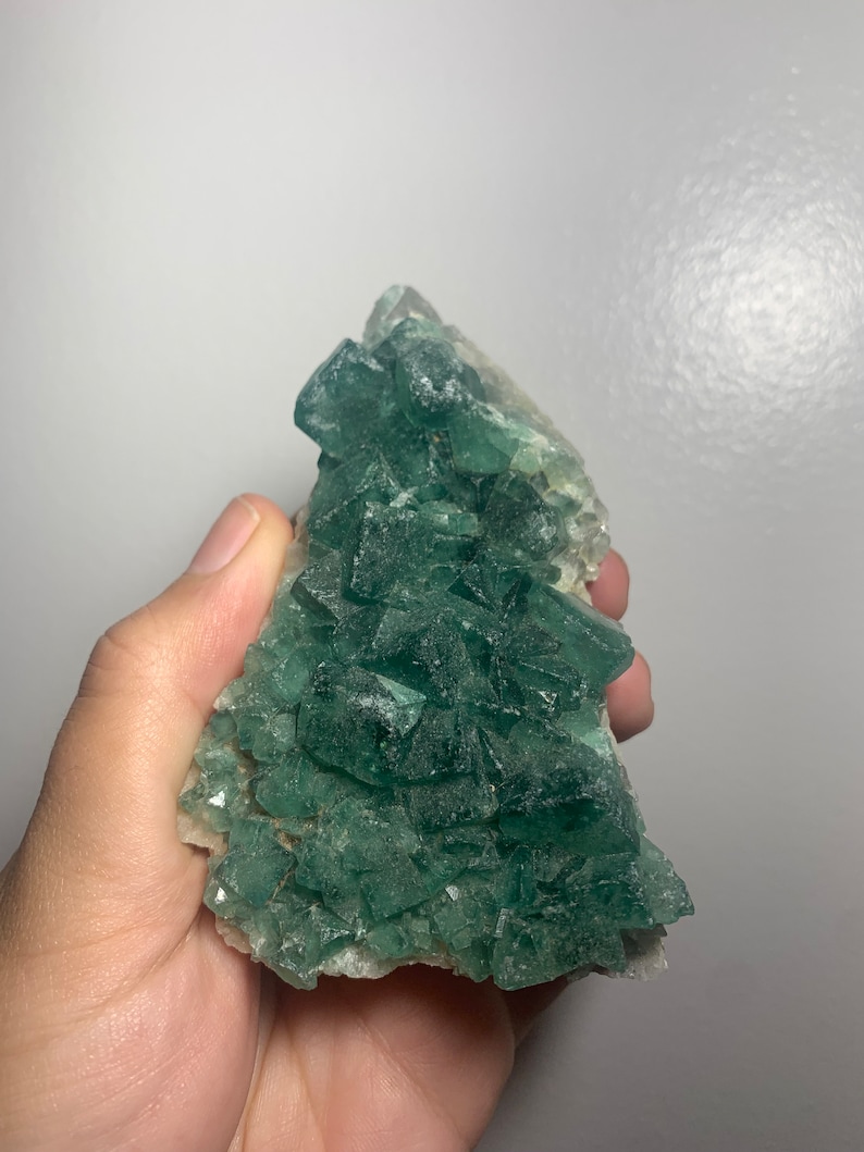 May include: A cluster of green fluorite crystals with a rough, natural texture. The crystals are arranged in a pyramid shape and have a translucent appearance.