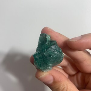 May include: A green fluorite crystal with a rough, natural texture. The crystal is a deep green colour with some lighter green areas. It is a beautiful example of a natural mineral.