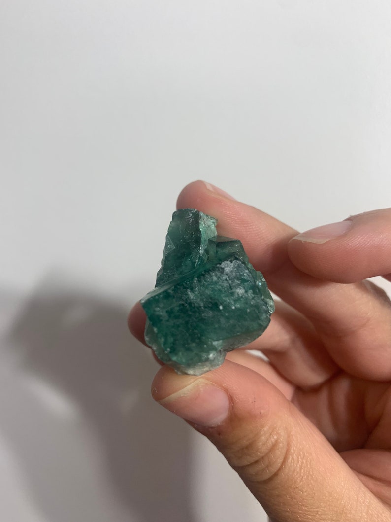 May include: A green fluorite crystal with a rough, natural texture. The crystal is a deep green colour with some lighter green areas. It is a beautiful example of a natural mineral.